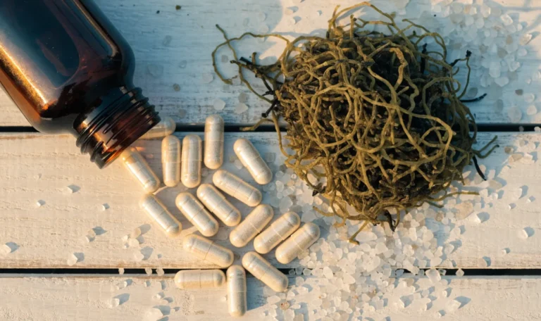 An open amber glass bottle spilling plain white plant-based capsules beside a small pile of dried wildcrafted sea moss, illustrating alkaline herb shop sea moss & moringa supplement 90 capsules.
