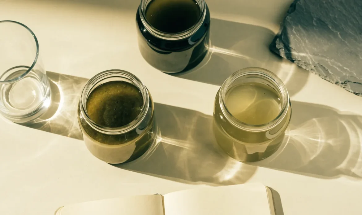 An unbranded flat lay comparing three plain glass jars of sea moss gel of different shades on a pale cream surface with a notebook, illustrating black girl vitamins sea moss.