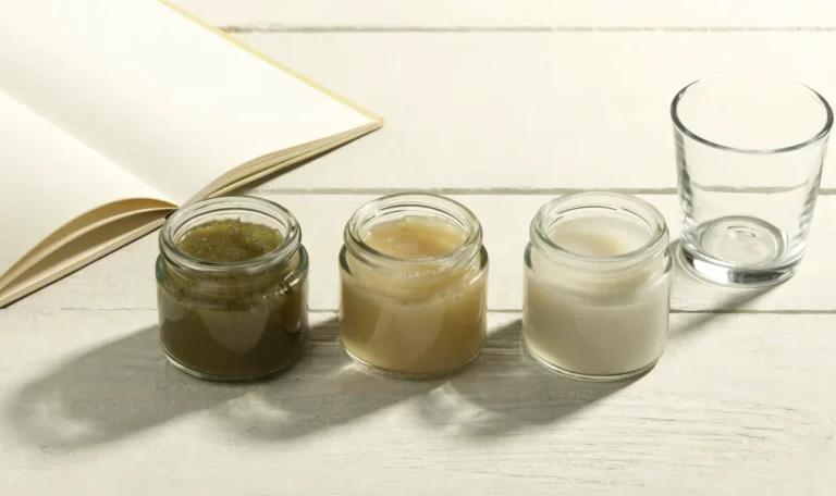 An unbranded flat lay comparing three plain glass jars of sea moss gel of different shades on a pale cream surface with a notebook, illustrating black owned sea moss.
