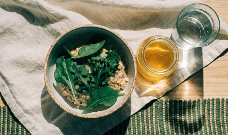 A simple bowl of oats and greens with a small jar of golden gel and a glass of water on a light wooden table, illustrating can i take sea moss on an empty stomach.