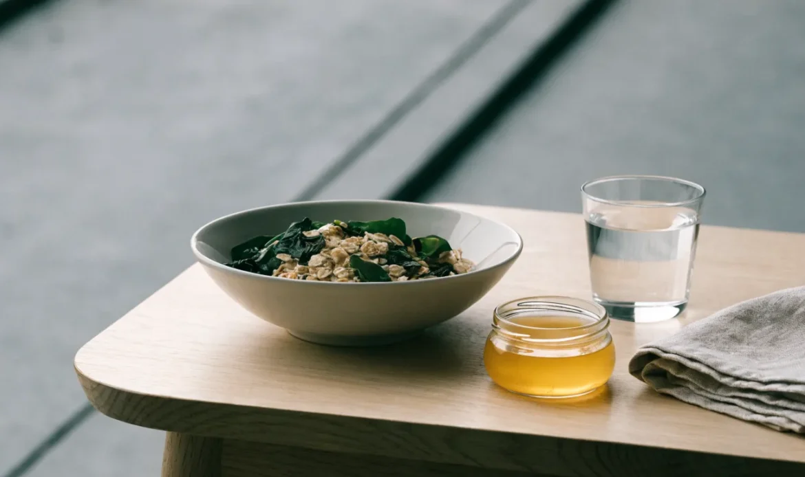 A simple bowl of oats and greens with a small jar of golden gel and a glass of water on a light wooden table, illustrating can sea moss make you sick.