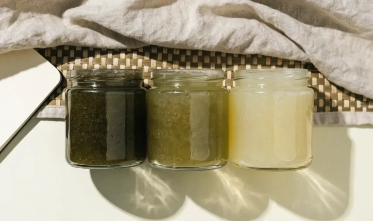 An unbranded flat lay comparing three plain glass jars of sea moss gel of different shades on a pale cream surface with a notebook, illustrating cgi green sea moss.