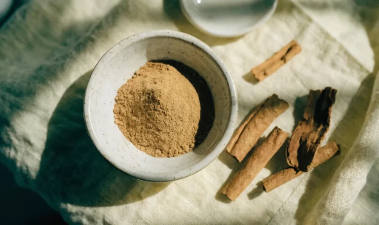 A small ceramic bowl of finely ground cinnamon powder beside broken cinnamon quills on a pale linen cloth, illustrating cinnamon cinnamon powder.