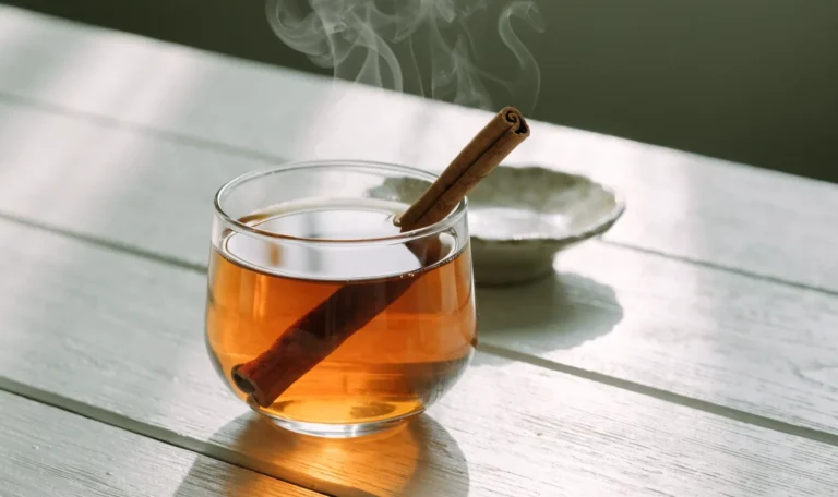A clear glass cup of amber spiced tea with a cinnamon quill resting inside, steam rising, on a wooden table, illustrating cinnamon stick for tea.