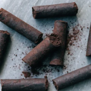 Roasted cocoa sticks on pale grey stone, hand-rolled cylinders of ground cacao with one snapped open to show the fudgy interior