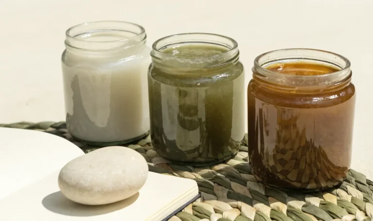 An unbranded flat lay comparing three plain glass jars of sea moss gel of different shades on a pale cream surface with a notebook, illustrating coconut cult and sea moss.