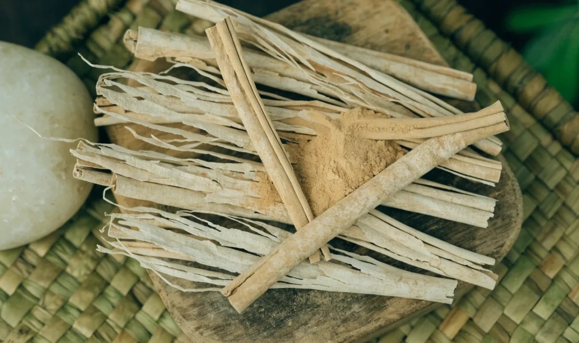 A loose bundle of pale, fragile, many-layered Ceylon cinnamon quills on a rustic wooden board with a small heap of ground cinnamon, illustrating crylon cinnamon.