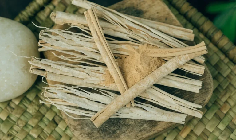 A loose bundle of pale, fragile, many-layered Ceylon cinnamon quills on a rustic wooden board with a small heap of ground cinnamon, illustrating crylon cinnamon.