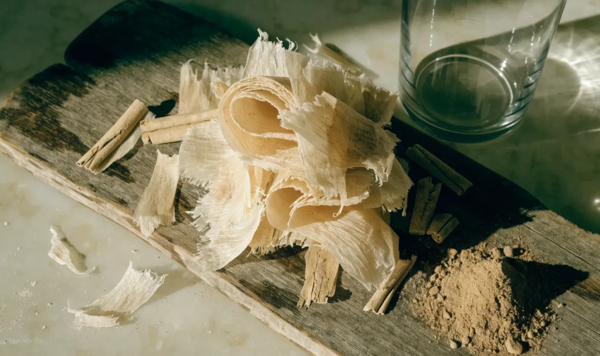 A loose bundle of pale, fragile, many-layered Ceylon cinnamon quills on a rustic wooden board with a small heap of ground cinnamon, illustrating cylon cinnamon.