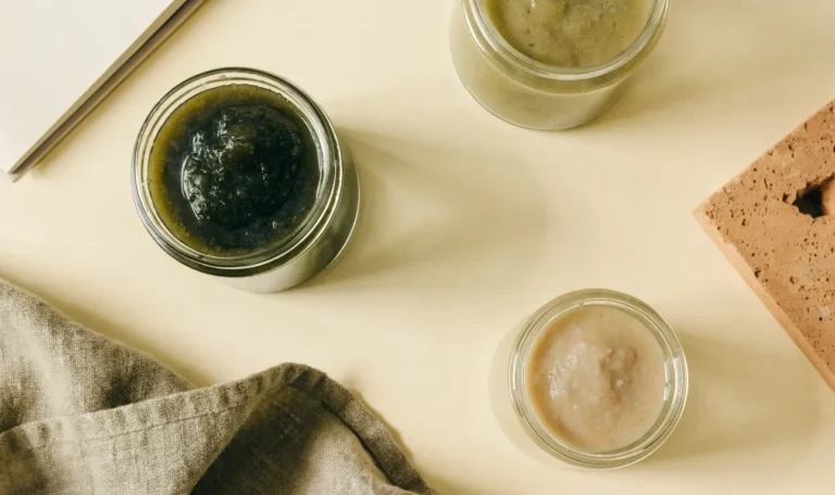 An unbranded flat lay comparing three plain glass jars of sea moss gel of different shades on a pale cream surface with a notebook, illustrating infinite age sea moss.