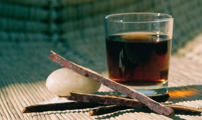Strips of dark reddish mauby tree bark on a woven mat beside a glass of dark amber bittersweet Caribbean bark drink, illustrating mauby bark.