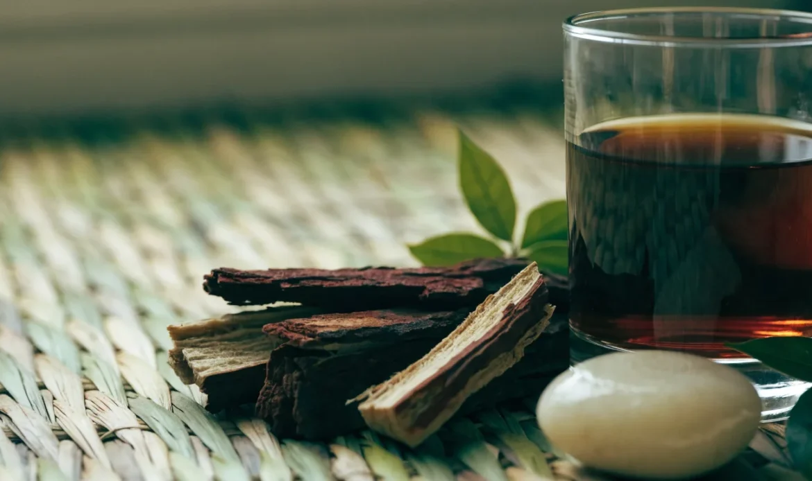 Strips of dark reddish mauby tree bark on a woven mat beside a glass of dark amber bittersweet Caribbean bark drink, illustrating mauby drink.