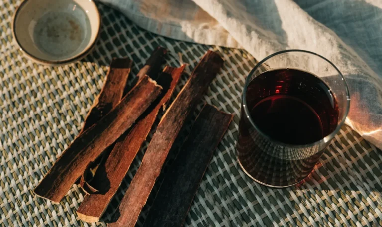 Strips of dark reddish mauby tree bark on a woven mat beside a glass of dark amber bittersweet Caribbean bark drink, illustrating mauby trinidad.