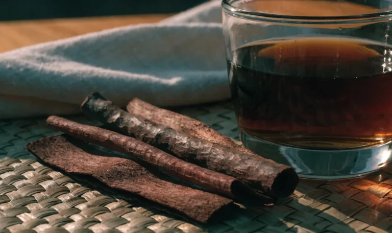 Strips of dark reddish mauby tree bark on a woven mat beside a glass of dark amber bittersweet Caribbean bark drink, illustrating mavi tree.