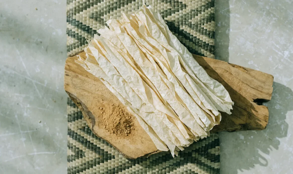 A loose bundle of pale, fragile, many-layered Ceylon cinnamon quills on a rustic wooden board with a small heap of ground cinnamon, illustrating salem cinnamon.