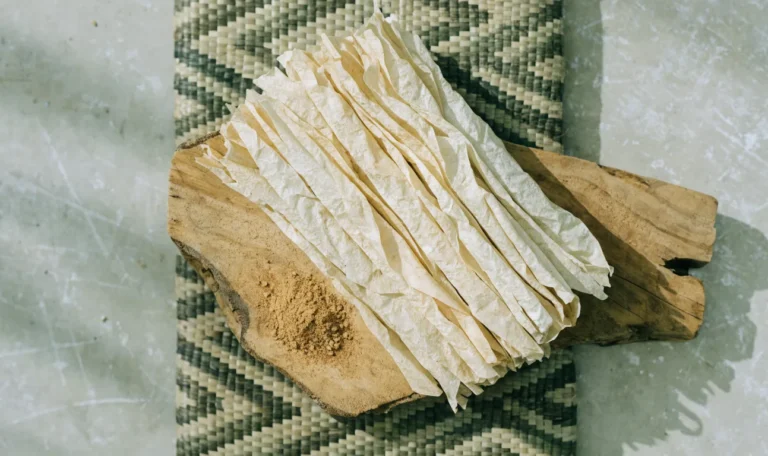 A loose bundle of pale, fragile, many-layered Ceylon cinnamon quills on a rustic wooden board with a small heap of ground cinnamon, illustrating salem cinnamon.