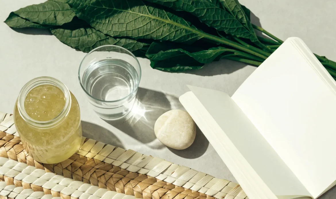 A clean flat lay of a small glass jar of sea moss gel, a notebook, a glass of water and fresh greens on a soft grey surface, illustrating seamoss and hypothyroidism.