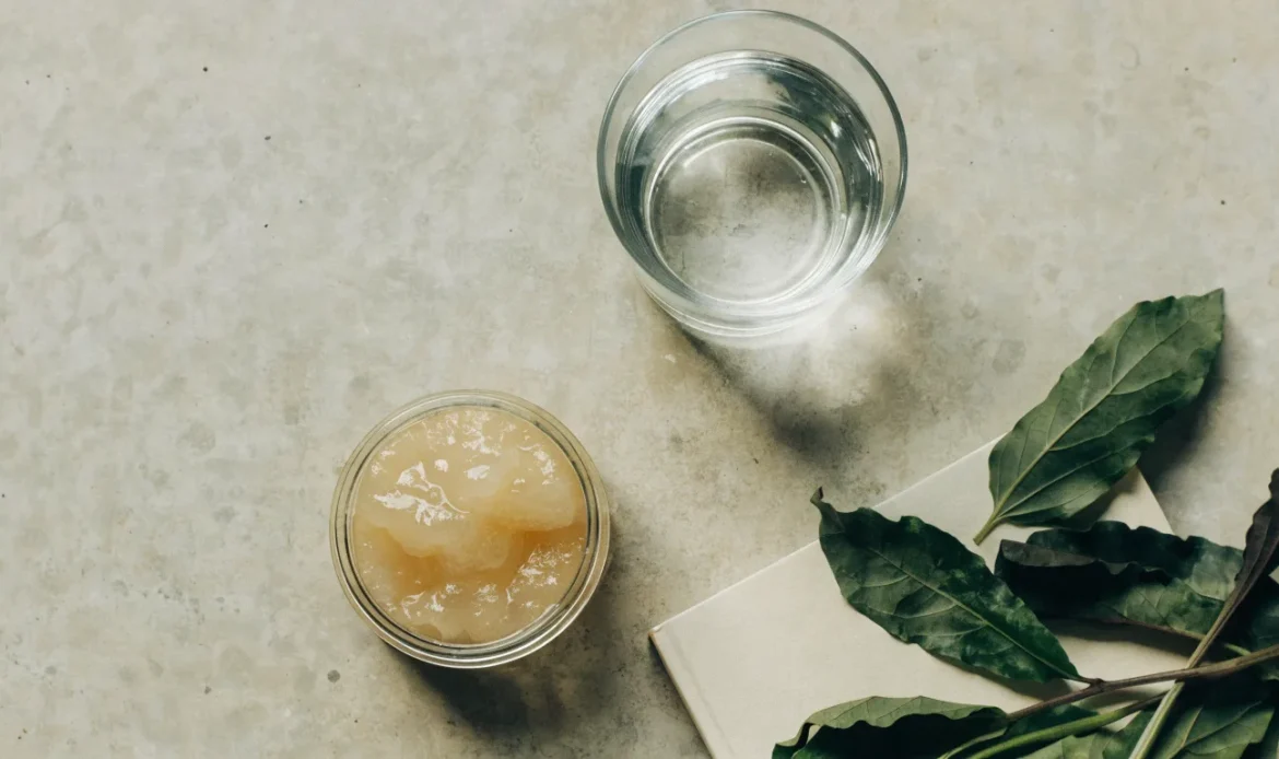 A clean flat lay of a small glass jar of sea moss gel, a notebook, a glass of water and fresh greens on a soft grey surface, illustrating sea moss for hormone balance.