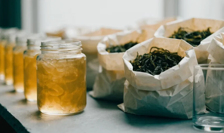 An unbranded market stall table of glass jars of golden gel and paper bags of dried seaweed under soft daylight, illustrating seamoss for sale.