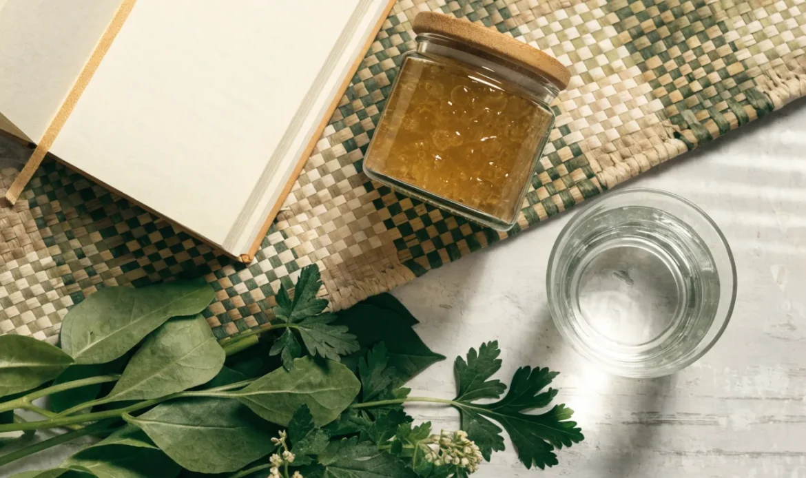 A clean flat lay of a small glass jar of sea moss gel, a notebook, a glass of water and fresh greens on a soft grey surface, illustrating sea moss for thyroid support.
