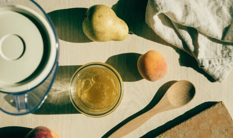 An overhead kitchen counter flat lay of a blender jug, a jar of golden gel, fresh fruit and a wooden spoon on light wood, illustrating sea moss recipes.