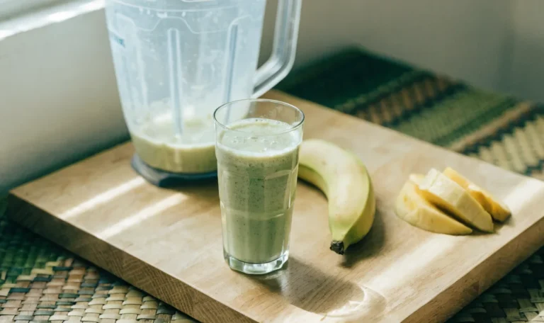 A tall glass of pale green smoothie on a light wooden counter with a blender jug, banana and mango slices behind, illustrating sea moss smoothie.