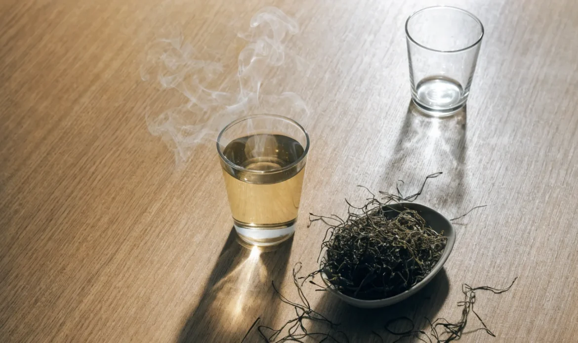 A clear glass cup of pale golden herbal infusion with steam rising, loose dried seaweed in a dish beside it, illustrating sea moss tea.