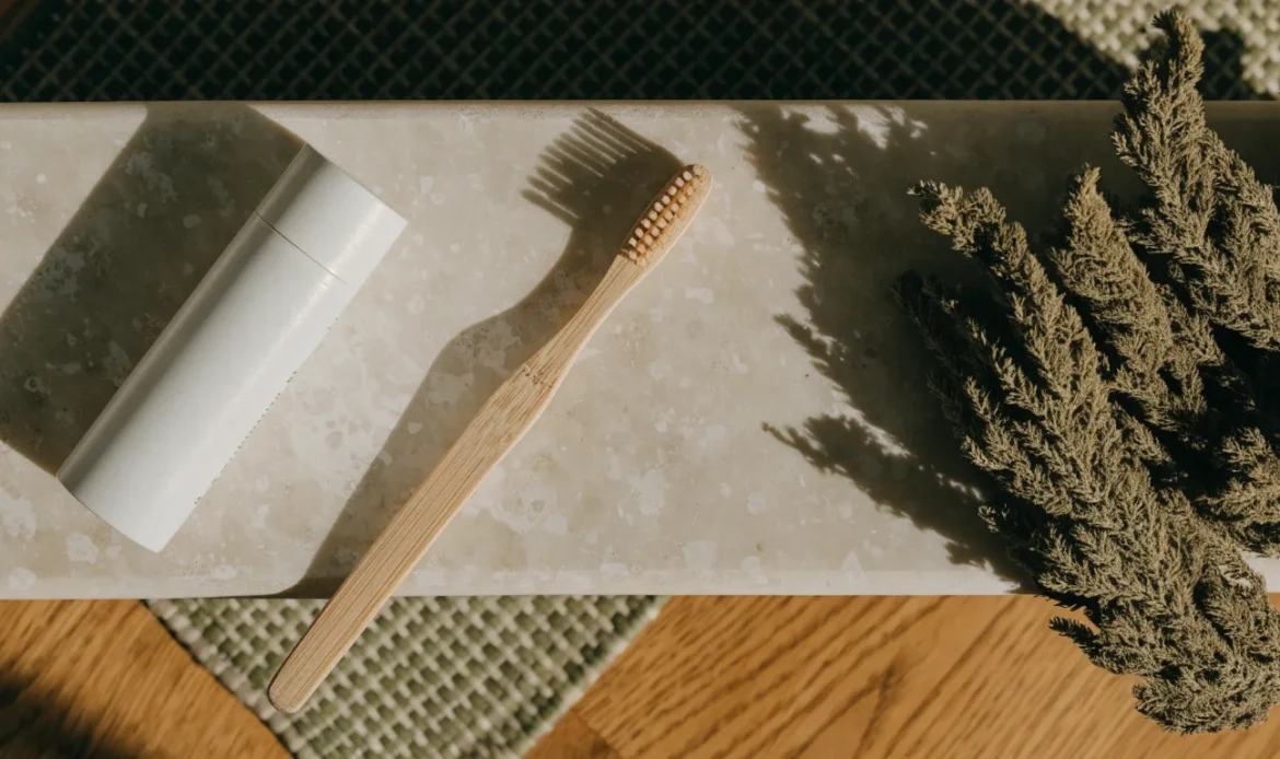 A plain white tube and a soft-bristle bamboo toothbrush on a pale stone bathroom shelf with a sprig of dried sea moss, illustrating sea moss toothpaste.