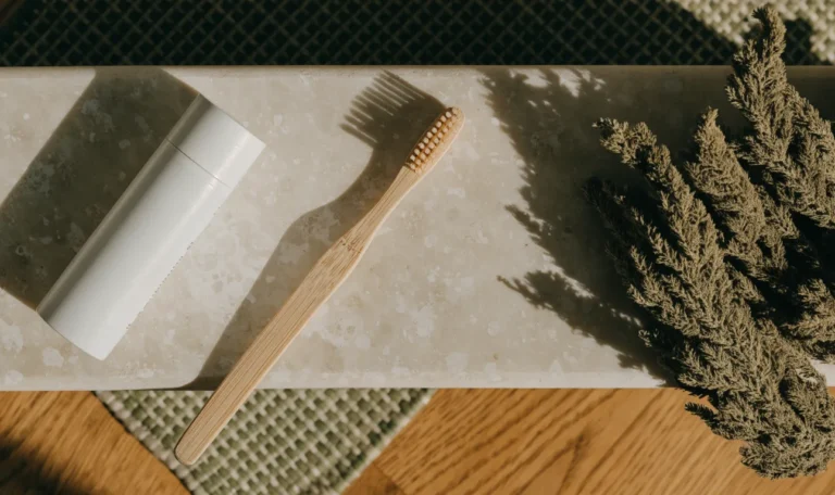 A plain white tube and a soft-bristle bamboo toothbrush on a pale stone bathroom shelf with a sprig of dried sea moss, illustrating sea moss toothpaste.