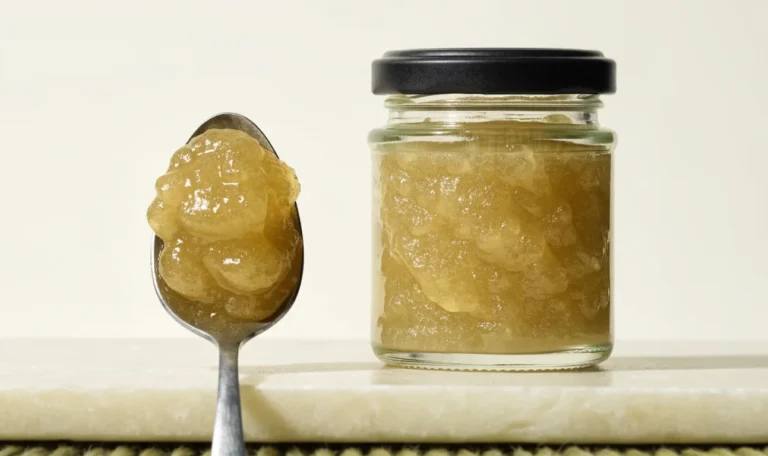 A small measured spoonful of golden sea moss gel beside a closed glass jar on a pale cream surface, restrained clinical styling, illustrating side effect of sea moss.
