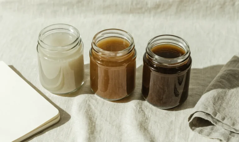 An unbranded flat lay comparing three plain glass jars of sea moss gel of different shades on a pale cream surface with a notebook, illustrating sim sima sea moss.