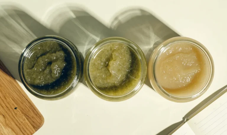 An unbranded flat lay comparing three plain glass jars of sea moss gel of different shades on a pale cream surface with a notebook, illustrating simsima sea moss.