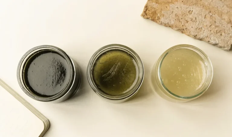 An unbranded flat lay comparing three plain glass jars of sea moss gel of different shades on a pale cream surface with a notebook, illustrating sunflower wellness sea moss.
