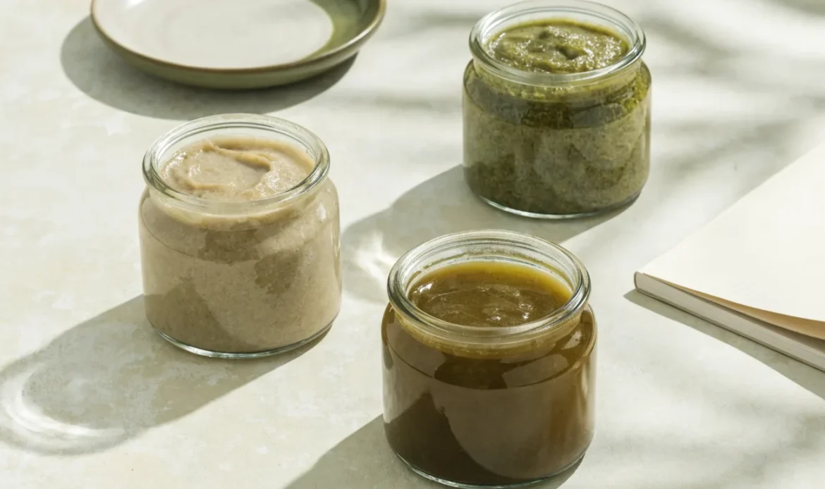 An unbranded flat lay comparing three plain glass jars of sea moss gel of different shades on a pale cream surface with a notebook, illustrating true sea moss reviews.