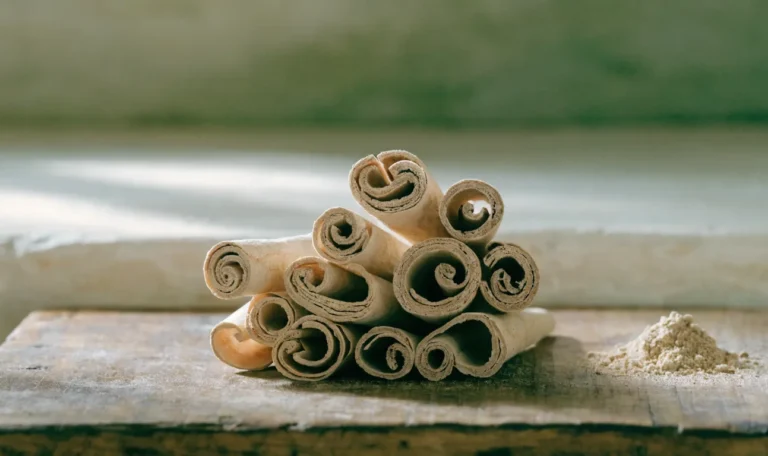 A loose bundle of pale, fragile, many-layered Ceylon cinnamon quills on a rustic wooden board with a small heap of ground cinnamon, illustrating where can i buy ceylon cinnamon near me.